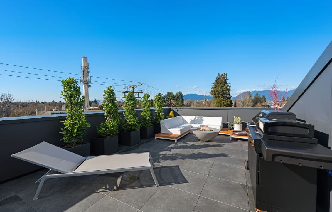 The Modern on Heather – A modern rooftop patio features dark tiled flooring, a sun lounger, a barbecue grill, and a white L-shaped outdoor sofa surrounding a circular concrete fire pit. Potted green trees line the dark parapet wall under a clear blue daylight sky, with distant mountains visible in the background.