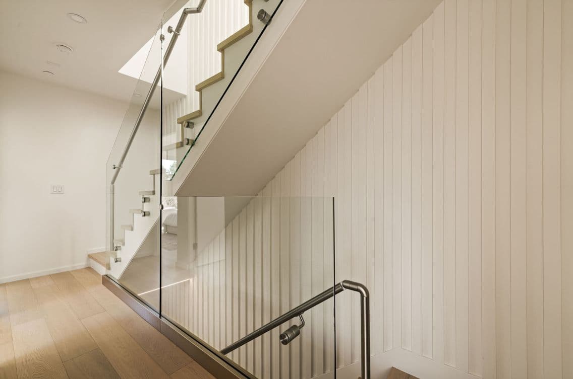 The Modern on Heather – A modern interior staircase features clear glass railings with stainless steel handrails set against light wood flooring and off-white vertical paneling. Soft recessed lighting overhead illuminates the clean lines and neutral palette of the multi-level home.