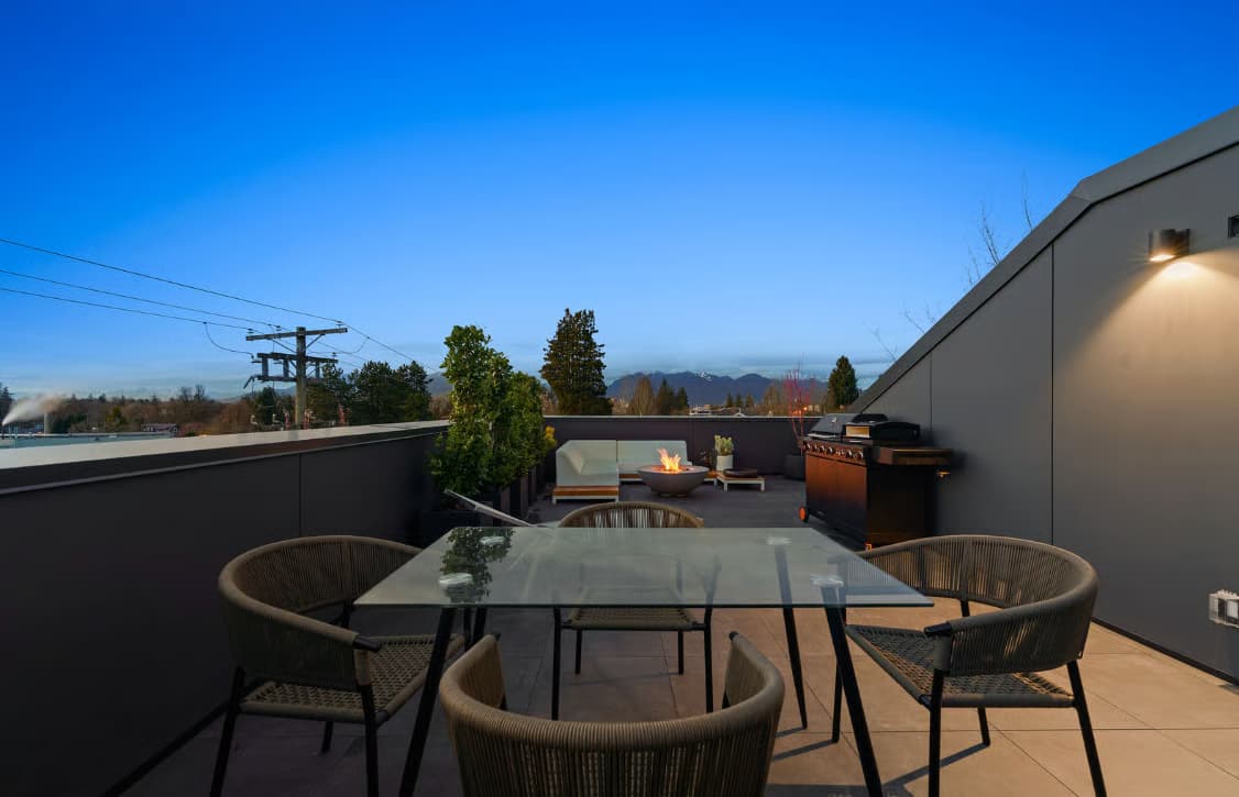 The Modern on Heather – A modern rooftop patio features a glass dining table surrounded by woven chairs in the foreground, with a barbecue grill, modular sectional lounge, and a lit fire bowl in the background. Set against a deep blue twilight sky, the space offers distant views of trees and mountain outlines.