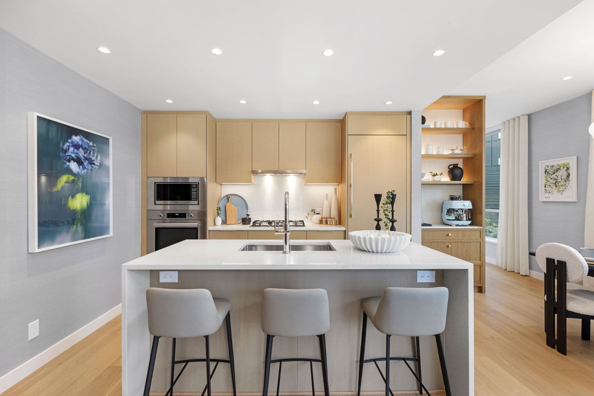 Exeter Hall – A modern, bright kitchen featuring light wood flat-panel cabinetry, integrated appliances, and a waterfall-edge white quartz island with three barstools. Recessed ceiling lighting illuminates the space, which includes an adjacent built-in shelving unit with a coffee station and light hardwood flooring.