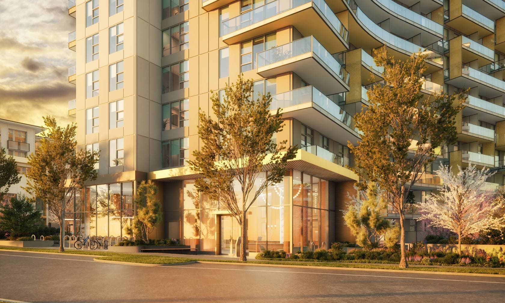 Exeter Hall – A ground-level view of a contemporary high-rise residential building featuring a double-height glass lobby entrance, street-level bike racks, and landscaped greenery. Glass-railed balconies curve along the upper facade, all illuminated by the warm, golden glow of late afternoon or sunrise light.