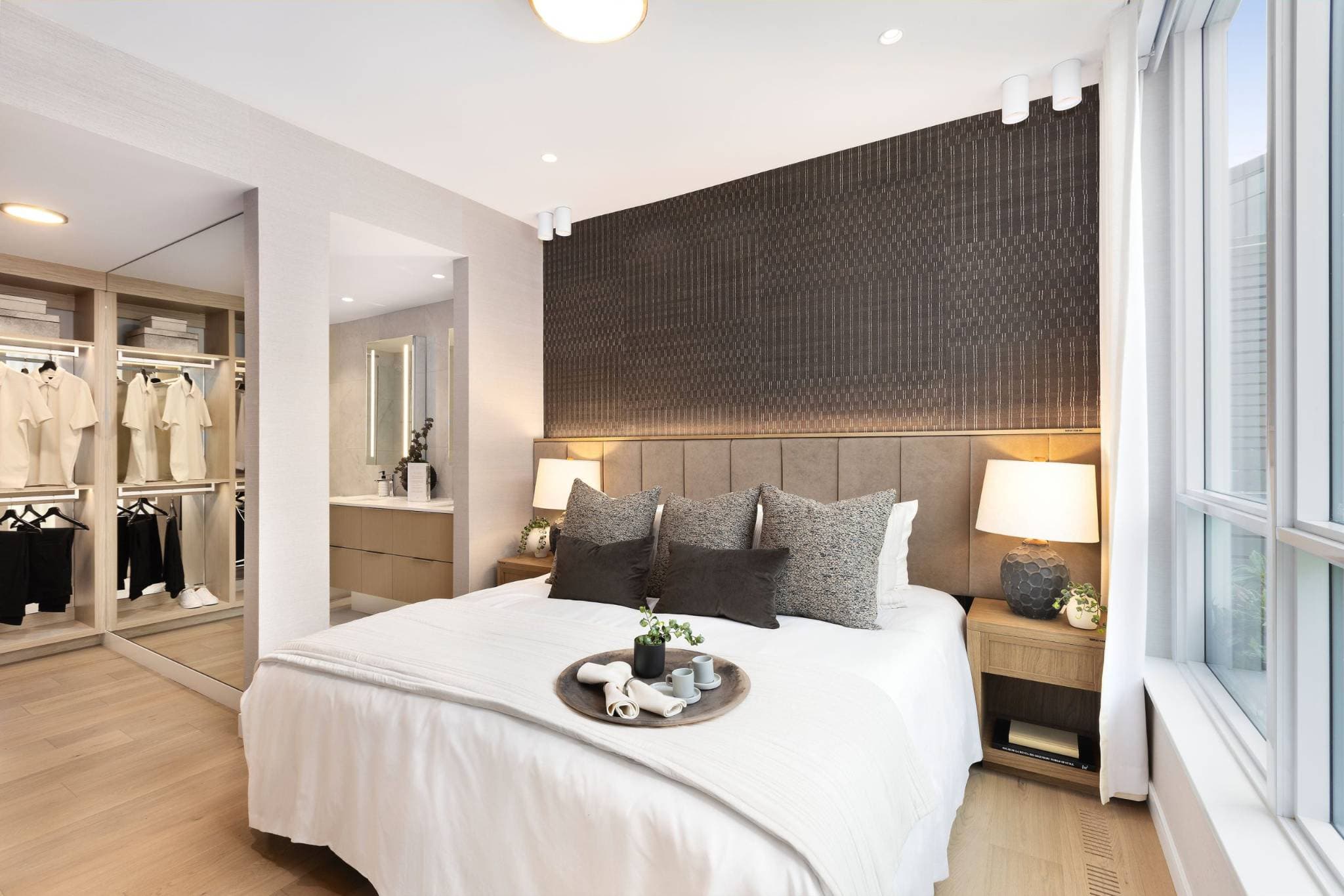 Exeter Hall – A modern bedroom suite features a made bed flanked by wooden nightstands with glowing table lamps against a dark, textured accent wall with integrated under-shelf lighting. To the left, the room flows into an illuminated walk-in closet and an ensuite bathroom vanity, all set over light wood floors alongside a large window.