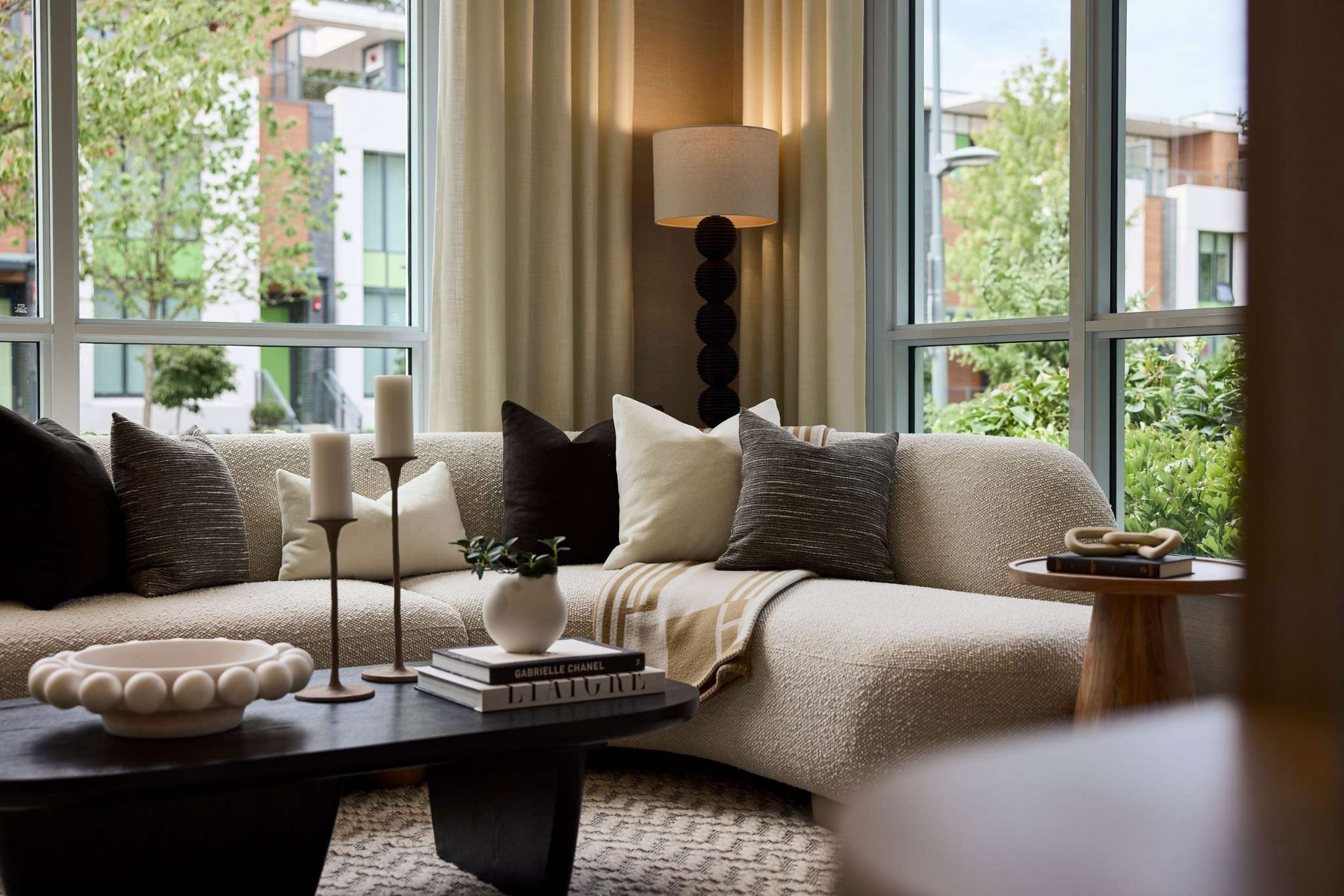 Exeter Hall – A bright living room corner features a curved, textured cream sectional sofa styled with accent pillows and a throw blanket, set against large floor-to-ceiling windows showing outdoor greenery. A low dark wood coffee table displays decorative candles and art books alongside a floor lamp tucked neatly in the corner.