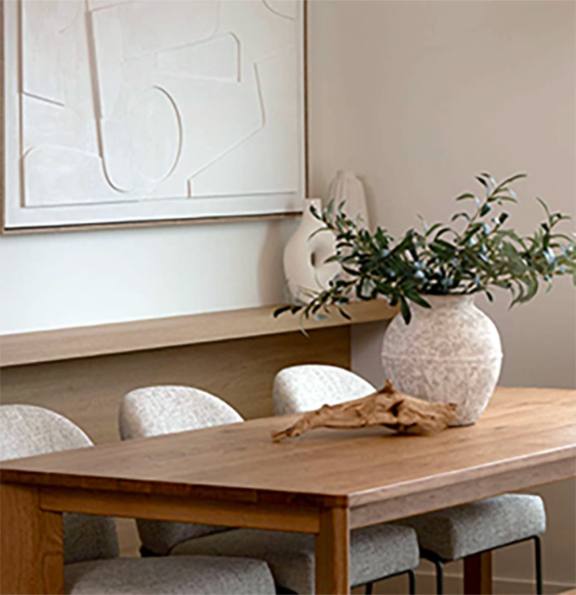 Heartwood – A warm and minimalist dining area featuring a natural wood dining table surrounded by textured grey upholstered chairs. The space is styled with an earthy ceramic vase holding leafy branches, a piece of driftwood, and a textured white abstract canvas hanging on the neutral wall above a wooden ledge.