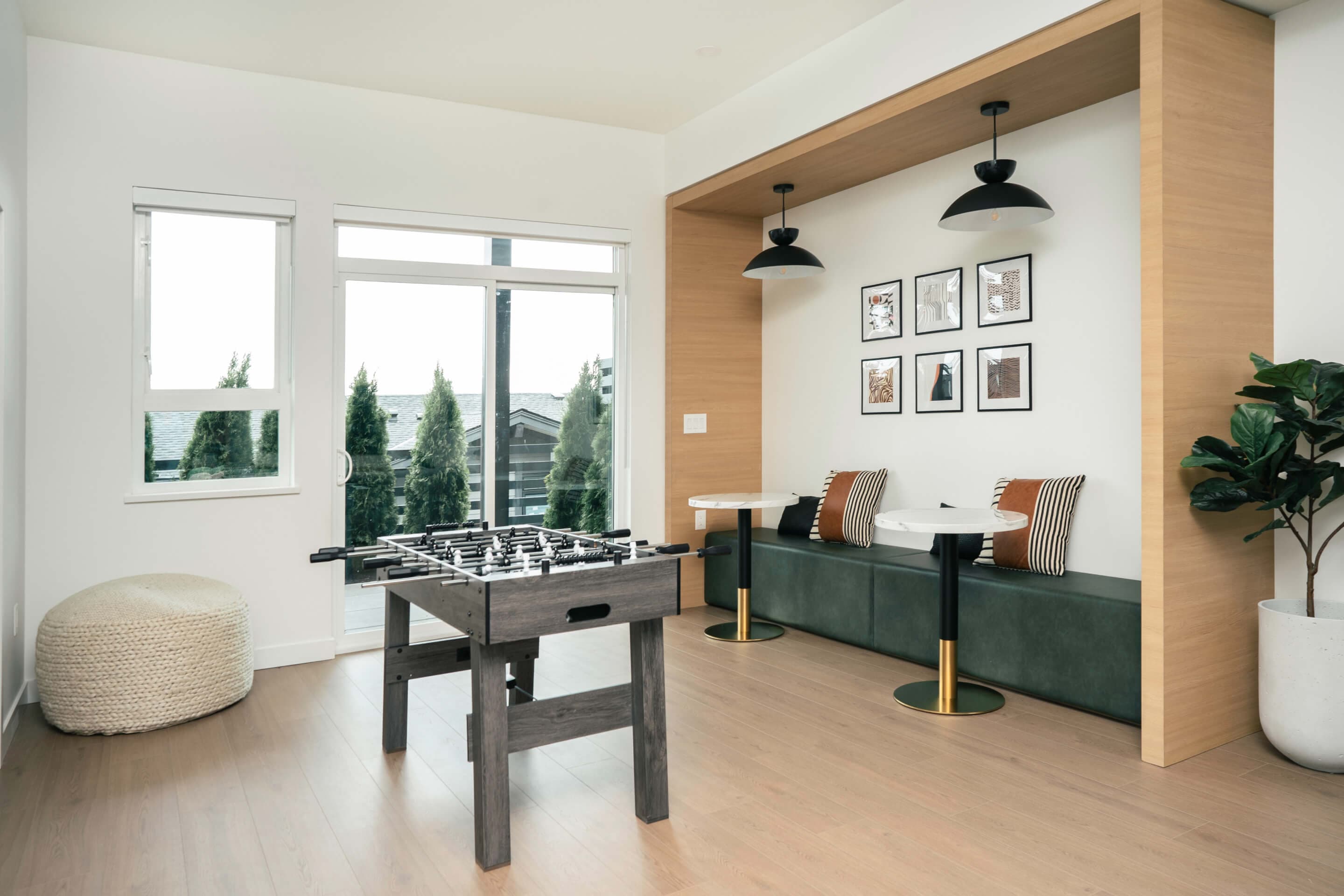 Heartwood – A modern games lounge featuring a weathered-wood foosball table, light hardwood floors, and sliding glass doors that open to an outdoor terrace with evergreens. A wood-framed seating nook contains a dark green bench, small marble café tables with brass bases, framed wall art, and black dome pendant lights.