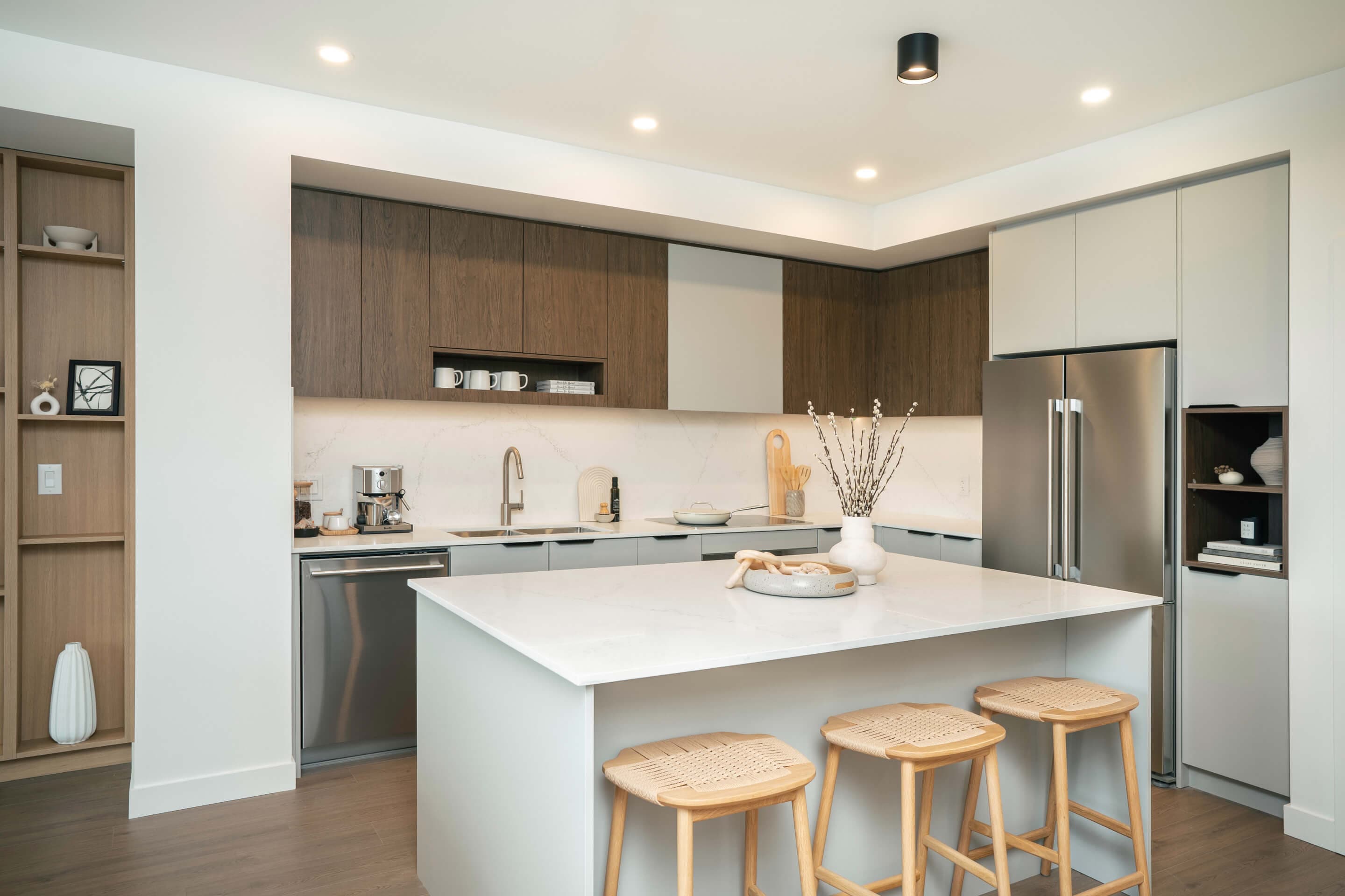 Heartwood – A modern open-concept kitchen features a central white quartz island paired with woven counter stools and recessed warm ceiling lighting. Flat-panel cabinetry in a mix of dark wood grain and soft grey surrounds stainless steel appliances and a white marble-patterned backsplash.
