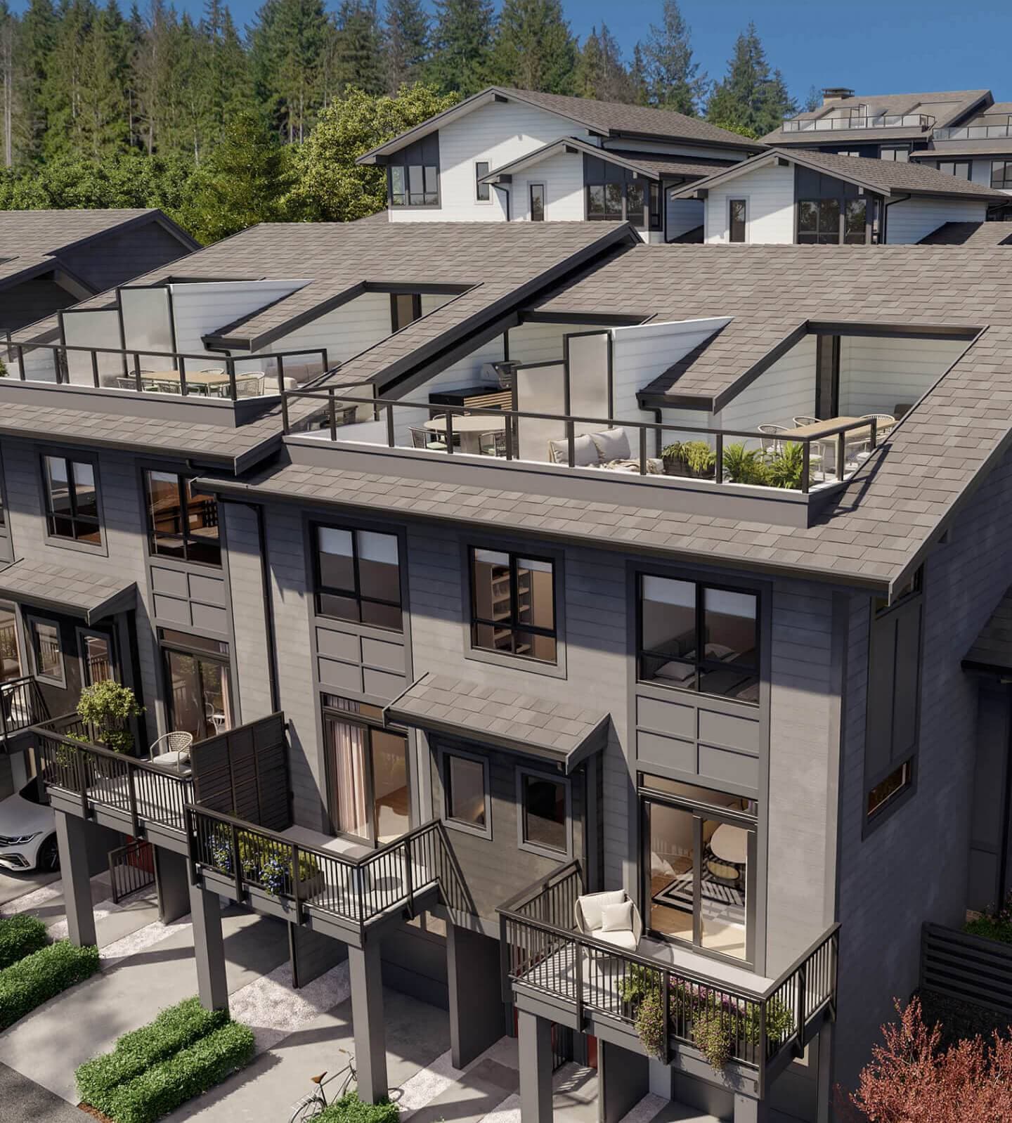 Heartwood – An elevated exterior view of modern multi-level townhomes featuring dark grey siding, individual balconies, and built-in rooftop terraces with outdoor dining and lounge areas. The contemporary homes are nestled into a hillside backdrop of lush evergreen trees under bright daylight.