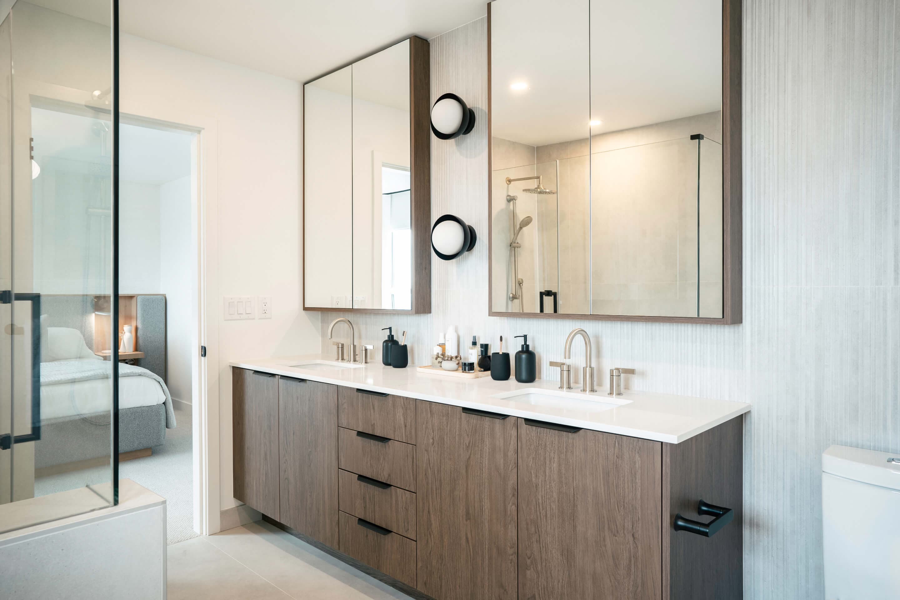 Heartwood – A modern ensuite bathroom features a double-sink vanity with dark wood grain cabinetry, a white countertop, and dual mirrored medicine cabinets with modern circular sconces. An open doorway offers a view into an adjacent bedroom, while reflections in the mirrors show a glass-enclosed shower with contemporary fixtures.