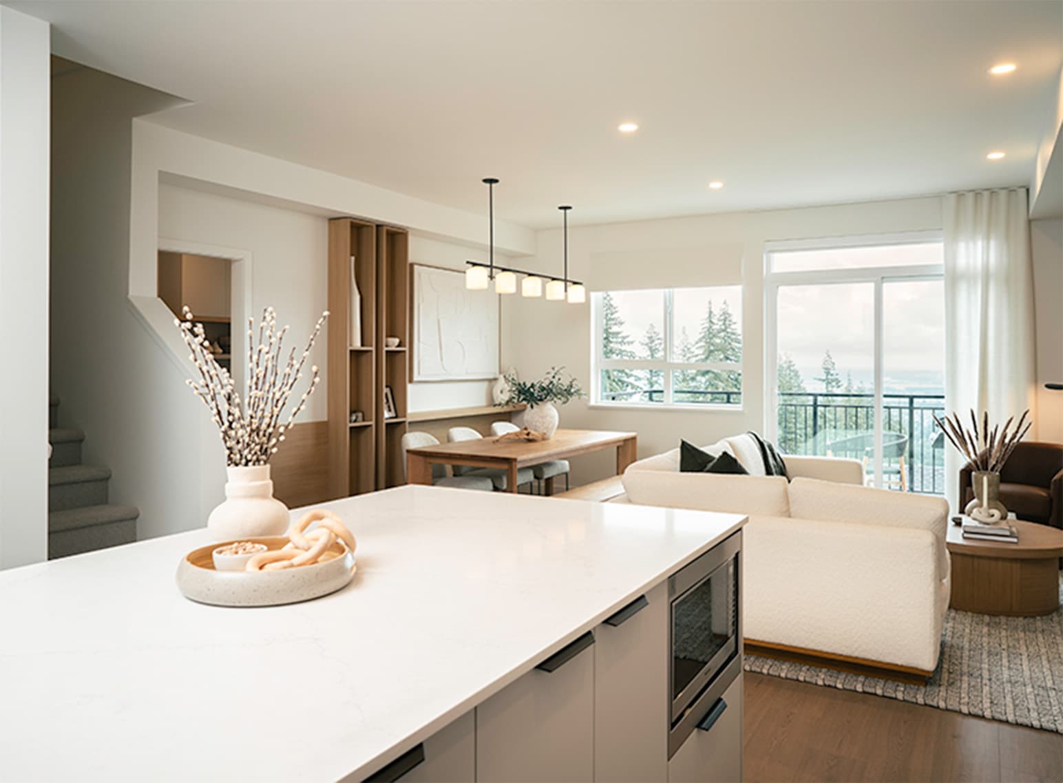 Heartwood – An open-concept living, dining, and kitchen area featuring a white quartz kitchen island in the foreground, light hardwood floors, and a modern dining table beneath linear pendant lighting. The adjacent living room includes a cream sofa and sliding glass doors that open to a balcony overlooking lush evergreen trees.