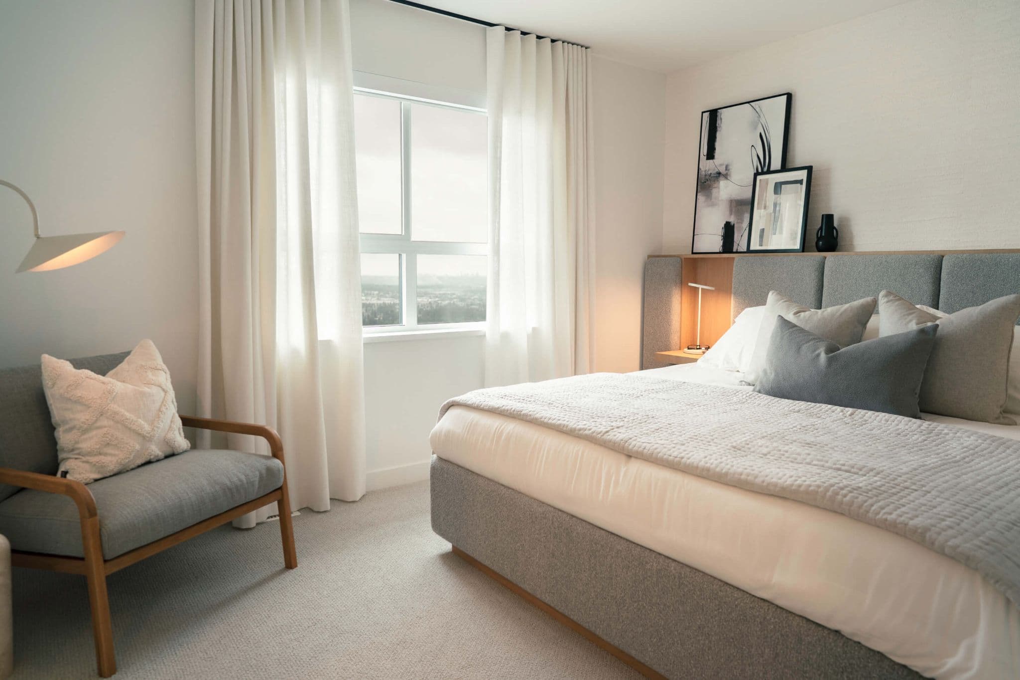 Heartwood – A bright, neutrally styled bedroom features an upholstered grey bed dressed in crisp white and grey linens, flanked by an integrated headboard with soft warm lighting. Floor-to-ceiling white drapes frame a window offering distant city and landscape views, alongside a cushioned wooden armchair and modern floor lamp.