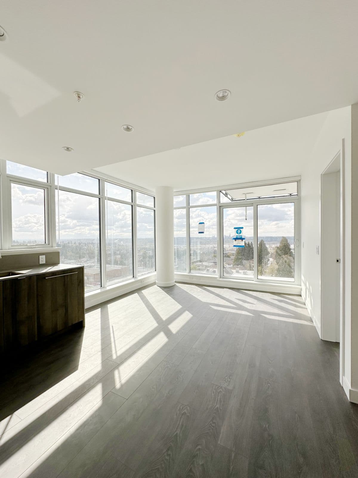 West – Empty living room corner with floor-to-ceiling windows showing a wide city and mountain view, dark wood flooring, and a glimpse of the kitchen island.
