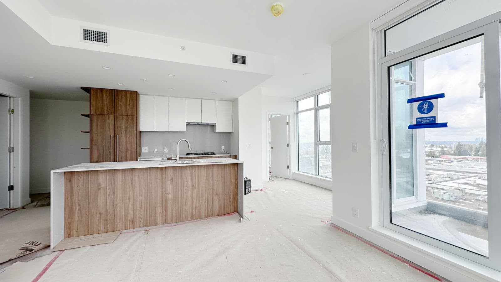 West – Kitchen under construction with a wood-fronted island, white and walnut cabinetry, protective floor covering, and a balcony door with a safety-harness sign.