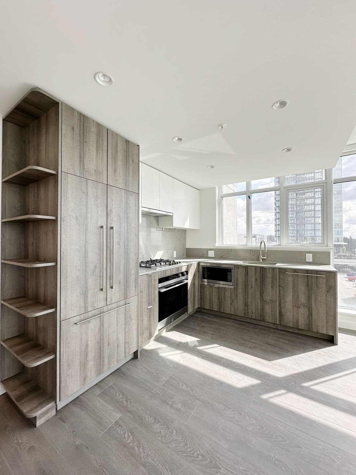 West – Finished kitchen with light wood cabinetry, built-in corner shelving, stainless appliances, and floor-to-ceiling windows overlooking the city skyline.