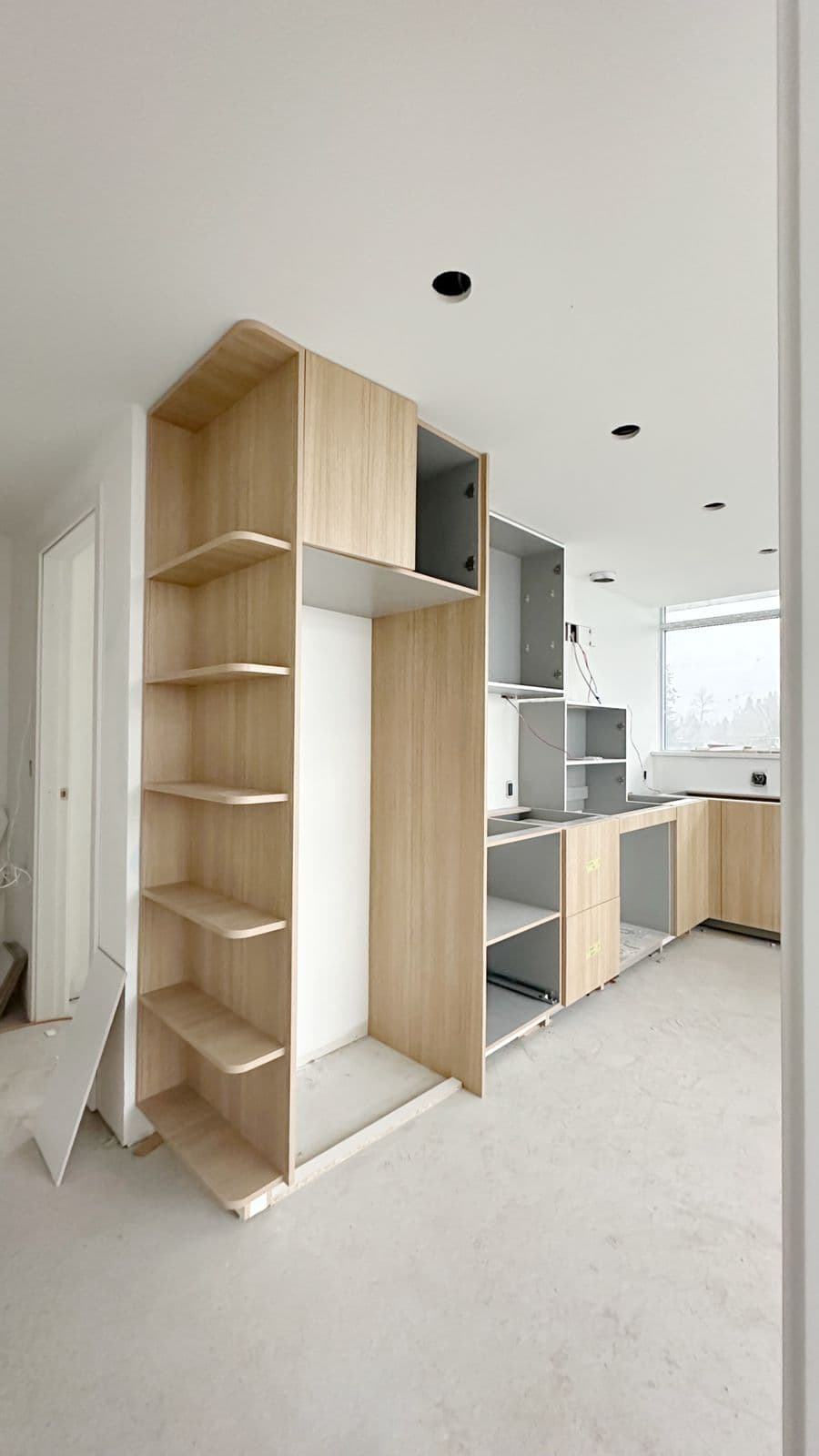 West – Built-in light wood cabinetry and open corner shelving under construction, with cabinet doors partially installed and a kitchen visible in the background.