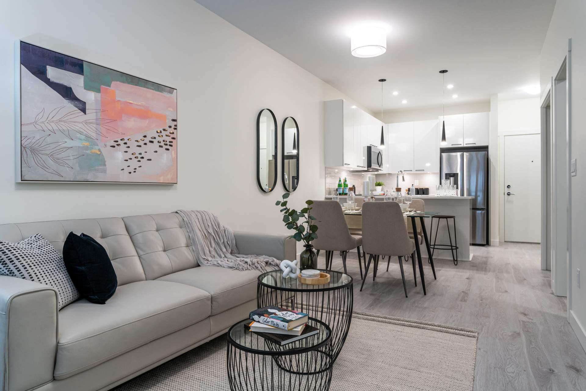 Sigmund – Rendering of an open living, dining, and kitchen space with a gray leather sofa, abstract artwork, a dining table, and a white kitchen with a stainless refrigerator.