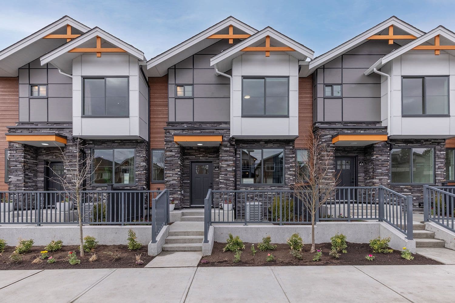 Harmony – A row of modern two-story townhomes viewed straight-on from across the sidewalk, featuring pitched gables with warm wood truss accents, dark stone masonry on the lower level, and grey panel siding above. Concrete steps lead up to individual gated front entryways bordered by grey metal railings and freshly landscaped mulched garden beds.