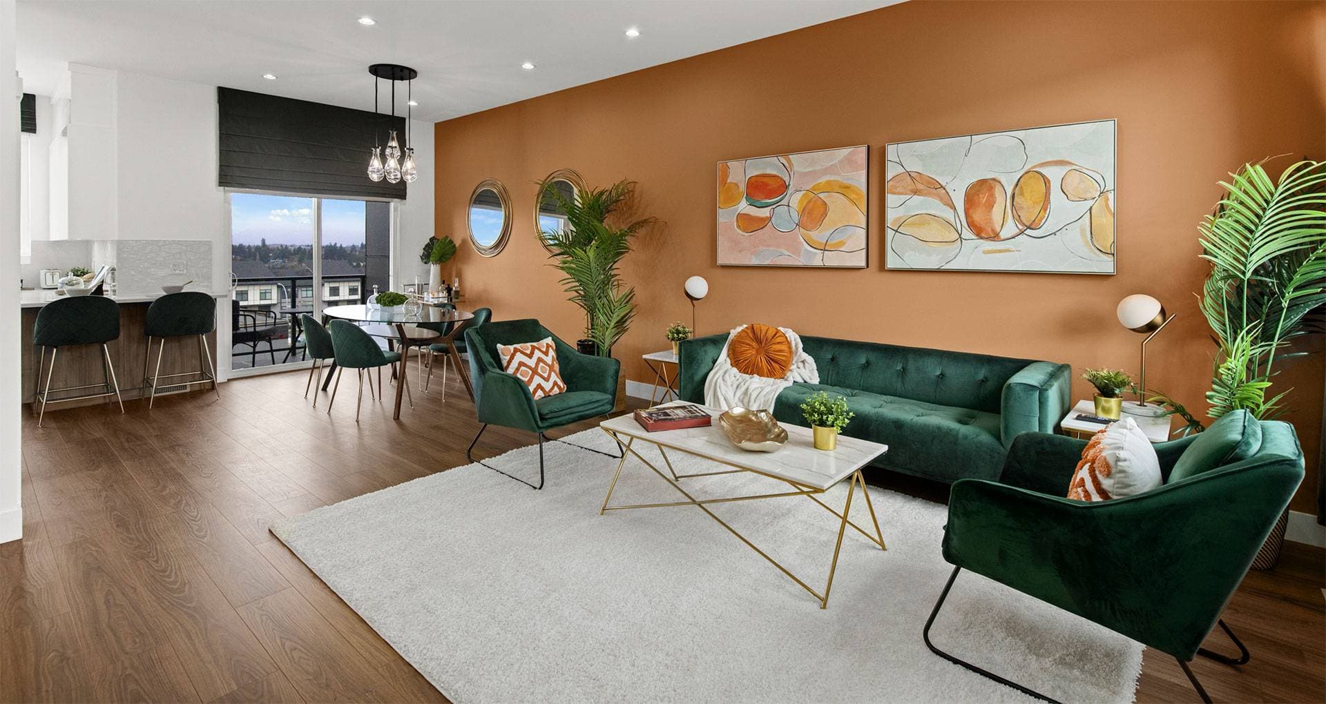 Harmony – An open-concept modern living space features a warm terracotta accent wall styled with abstract art and round mirrors above a green velvet sofa and matching armchairs. Wood flooring extends past a glass-topped dining set toward sliding doors that open onto a balcony, while a marble-and-gold coffee table sits centered on a light area rug.