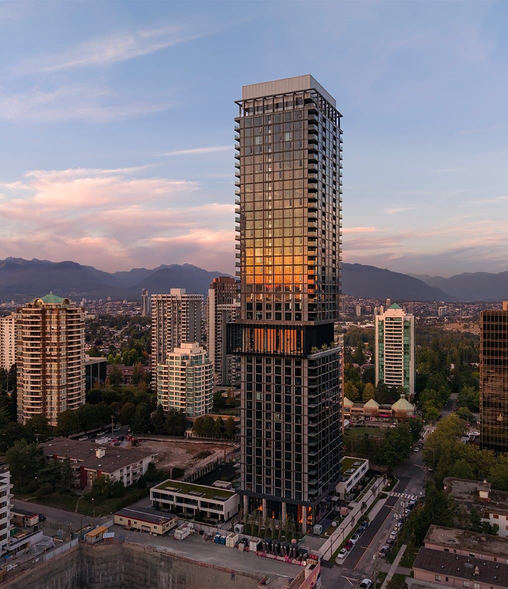 Central Park House – An elevated shot showcases a sleek, modern skyscraper featuring glass cladding that reflects a warm dusk sunset, rising above a dense urban residential area. Surrounding mid-rise buildings, green tree canopies, and distant mountain silhouettes frame the tower under a soft pink and blue sky.