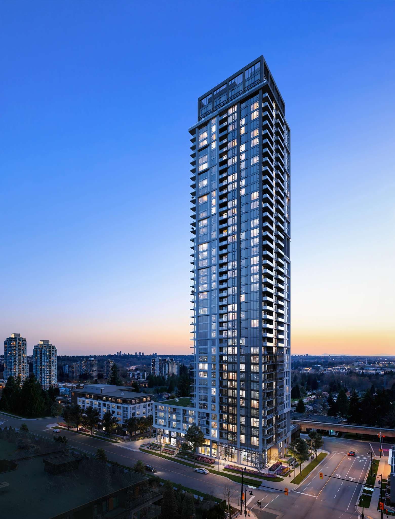 Vue – An elevated twilight shot of a slender, modern glass-and-steel residential skyscraper illuminated from within, rising above a podium along quiet city streets. The surrounding cityscape features low-rise buildings, mature green trees, and distant high-rises set against a gradient dusk sky.
