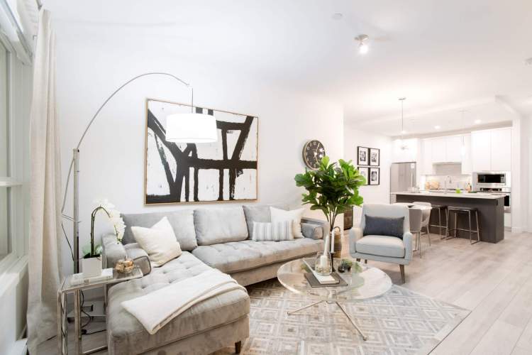 Highpointe – Open-concept living room with a grey tufted sectional sofa, black and white abstract art, and a view through to the white kitchen with an island and barstools.