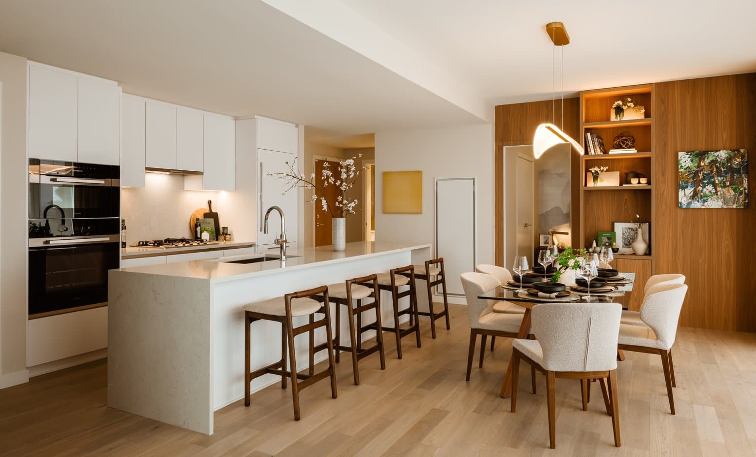 Claridge House – A modern open-concept kitchen and dining area features a large waterfall-edge island with bar seating, white flat-panel cabinetry, and integrated high-end appliances. Adjacent to the island, an elegant dining table is set with upholstered chairs beneath a sculptural pendant light, accented by warm wood wall paneling and built-in shelving.