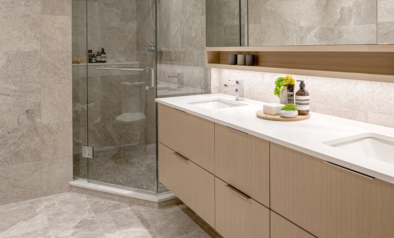 Claridge House – A modern bathroom interior featuring a floating light-wood double vanity with a white countertop and under-mirror accent lighting. In the background, a glass-enclosed walk-in shower is tiled in light gray stone that matches the surrounding walls and floor.