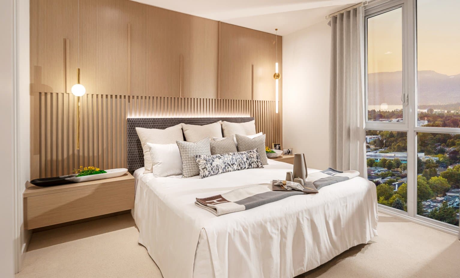 Claridge House – A modern bedroom featuring a bed dressed in neutral linens against a light-wood panel and slatted accent wall with integrated backlighting. Floor-to-ceiling corner windows look out over lush green neighborhoods toward distant mountains and water under a soft sunset sky.