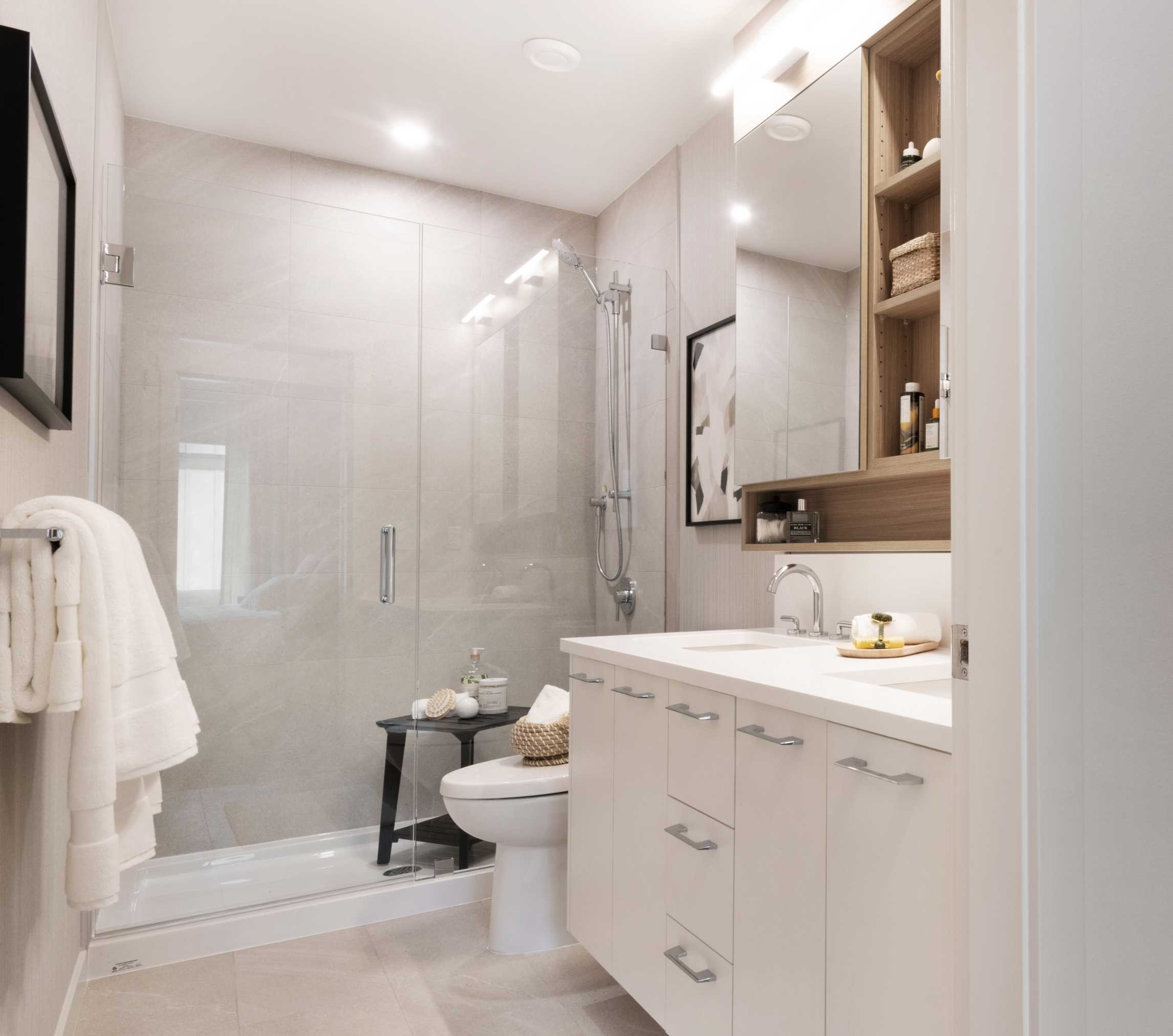 Cascadia – Bathroom with a glass-enclosed shower, toilet, and a white vanity with wood-accented shelving.