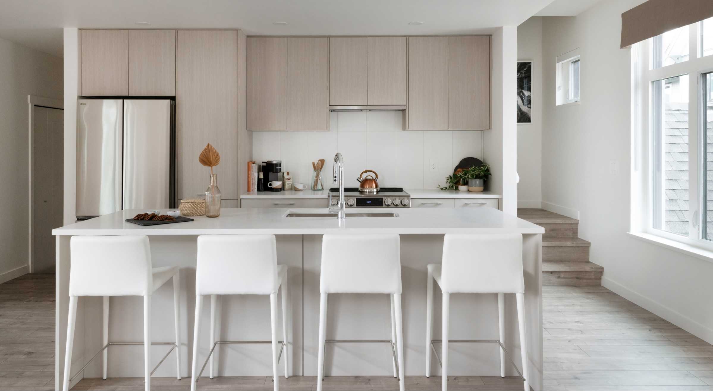 Cascadia – A kitchen island with white quartz countertop and four white bar stools, facing wood-toned cabinetry and a stainless steel fridge.