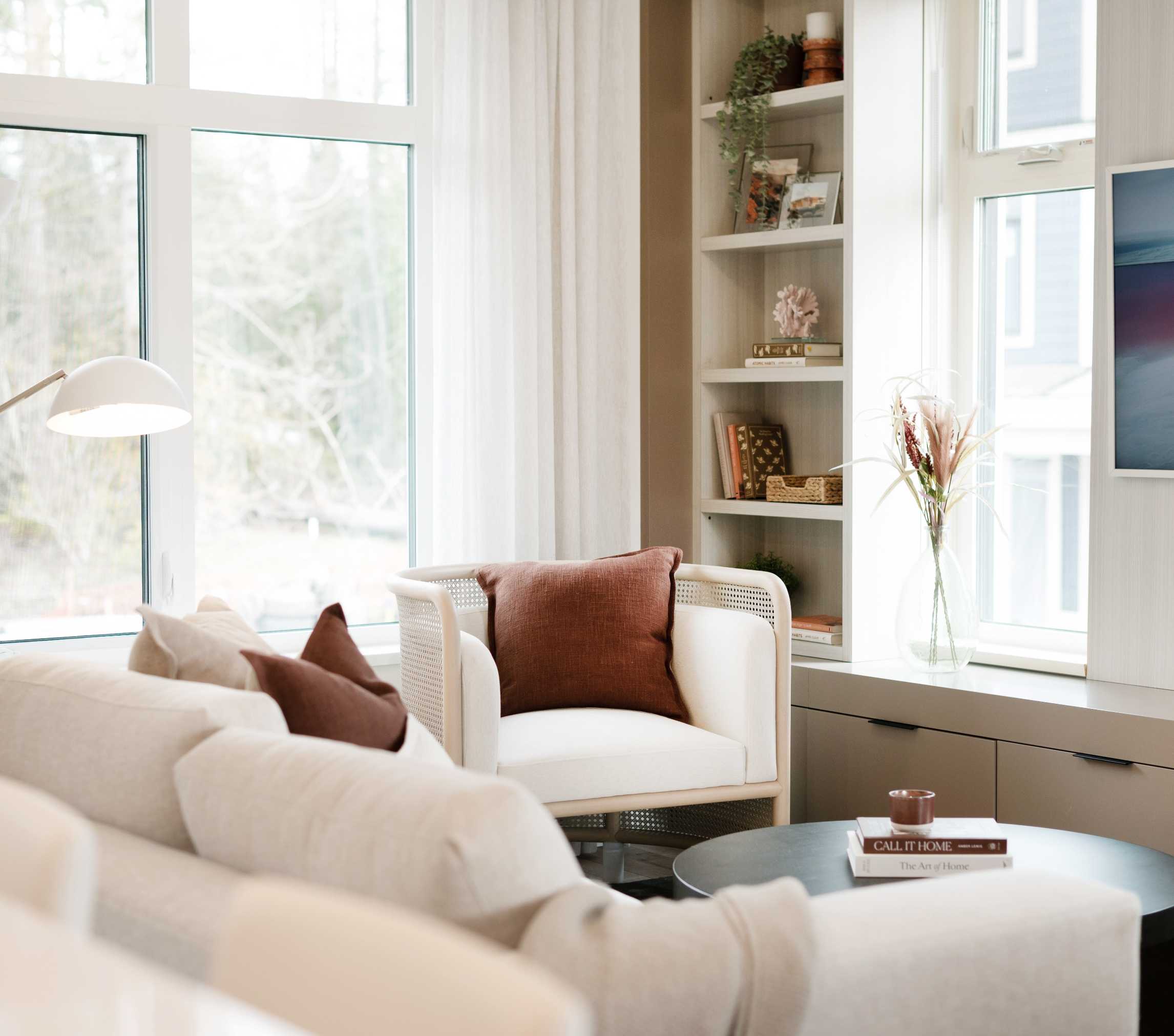 Cascadia – Living room corner with a cane-back armchair, built-in bookshelf, and beige sofas near large windows.