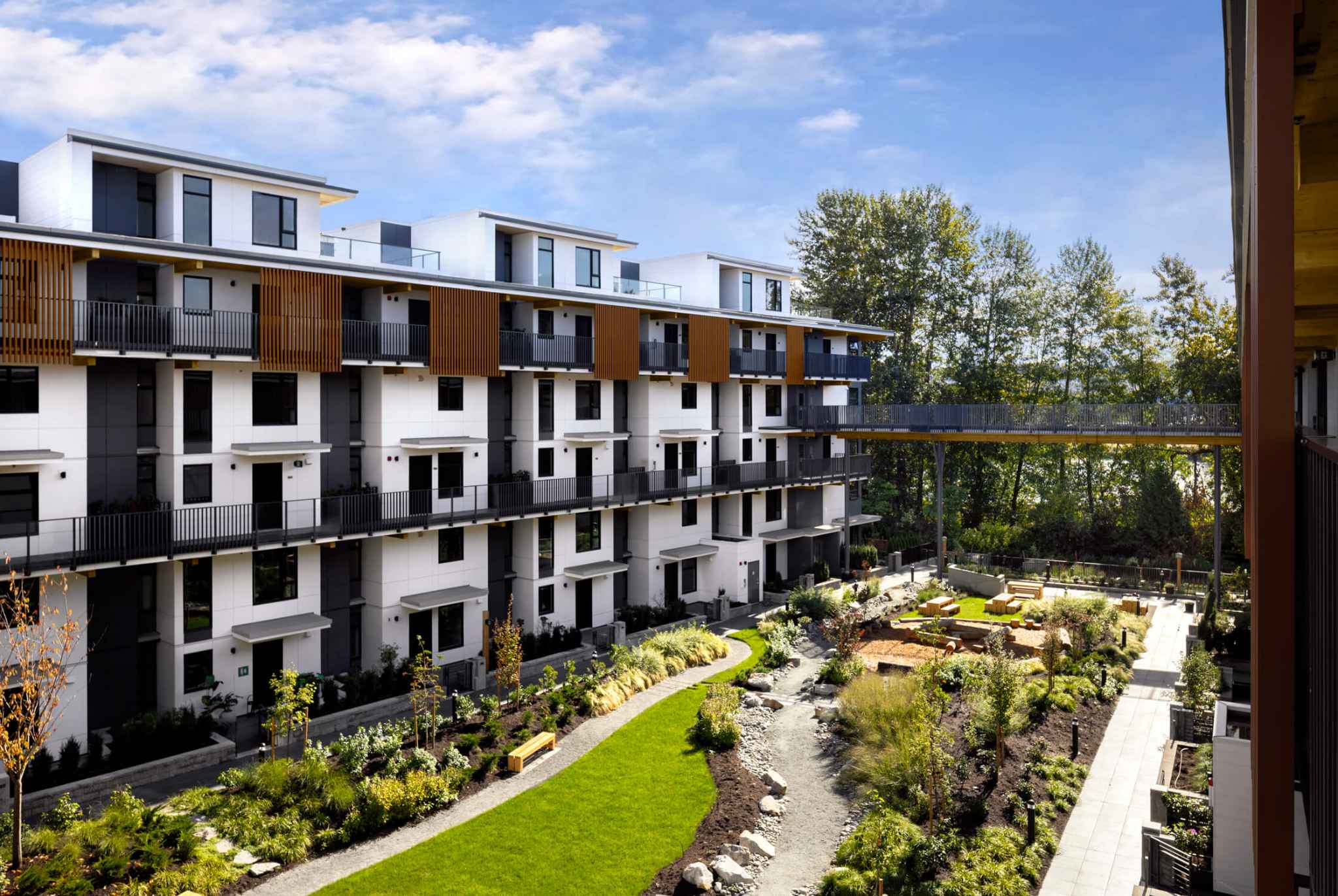 Timber House – Elevated view over the central landscaped courtyard: winding gravel creek bed, lawn, benches and a fire-pit seating area, with an elevated pedestrian bridge connecting the wood-accented residential wings and mature trees behind.