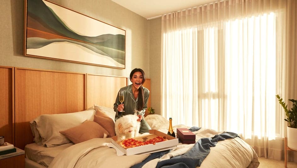 Parkside at Lynn – Bedroom with a woman and a dog on the bed sharing a pizza and champagne, a large landscape painting above the headboard, and sheer curtains at a bright window.