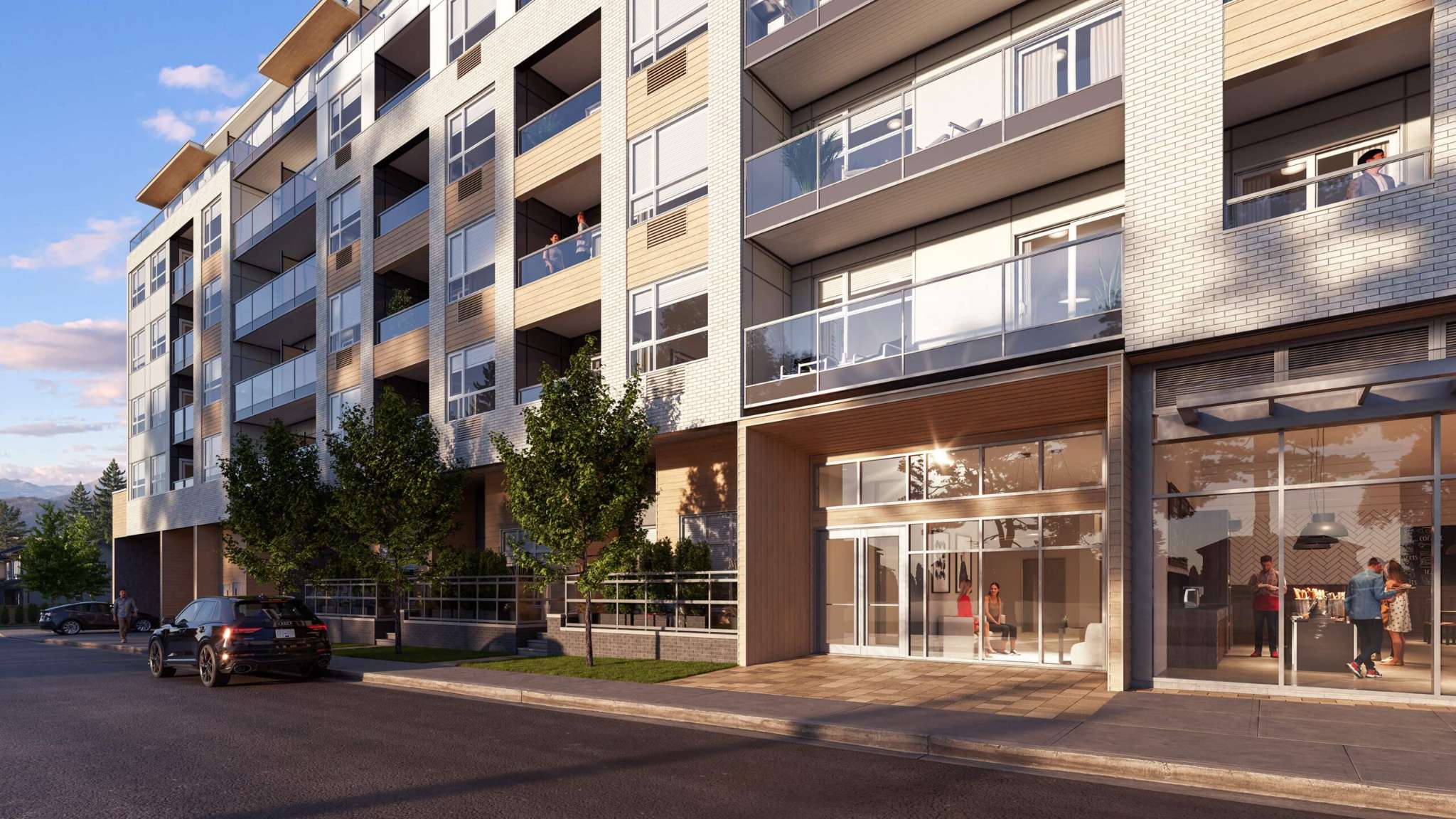 City and Laurel – A ground-level view of a contemporary mid-rise apartment building featuring glass-railed balconies, white brick and wood-panel accents, and ground-floor retail alongside a glass-enclosed residential entrance. A quiet street with a sidewalk, young street trees, and parked cars borders the facade under a clear late-afternoon sky.