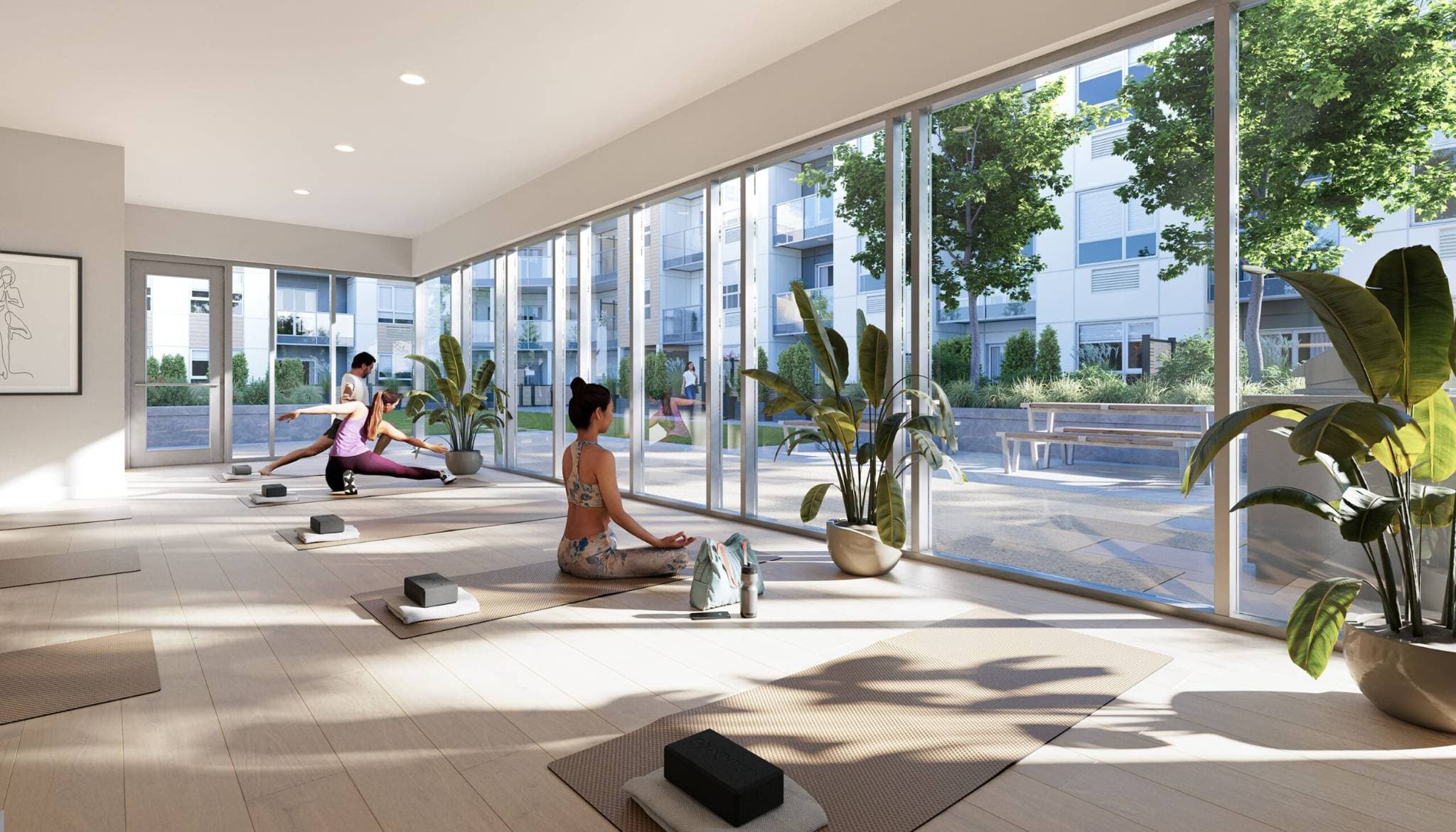 City and Laurel – A sunlit indoor yoga studio featuring light wood floors and large potted plants, with several people practicing yoga on mats facing floor-to-ceiling windows. Through the glass, a bright outdoor courtyard is visible, complete with paved walkways, green landscaping, picnic tables, and surrounding residential buildings.