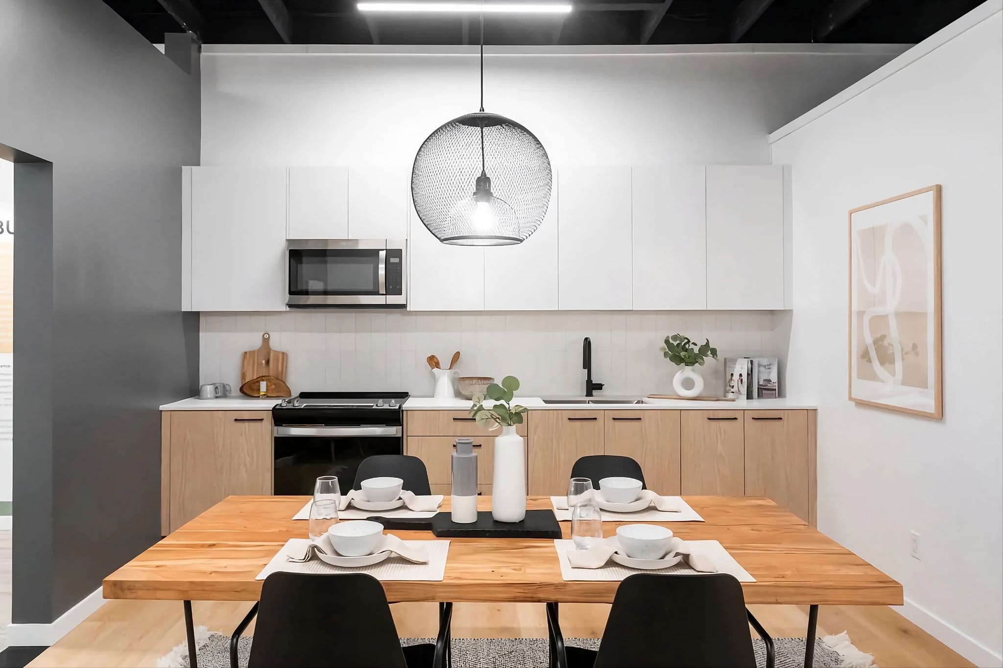 City and Laurel – A modern dining area and kitchen setup featuring a light-wood dining table set for four under a large black mesh globe pendant light. In the background, the kitchen includes two-tone cabinetry with light natural wood base cabinets, flat-panel white upper cabinets, stainless steel appliances, and white vertical tile backsplash.