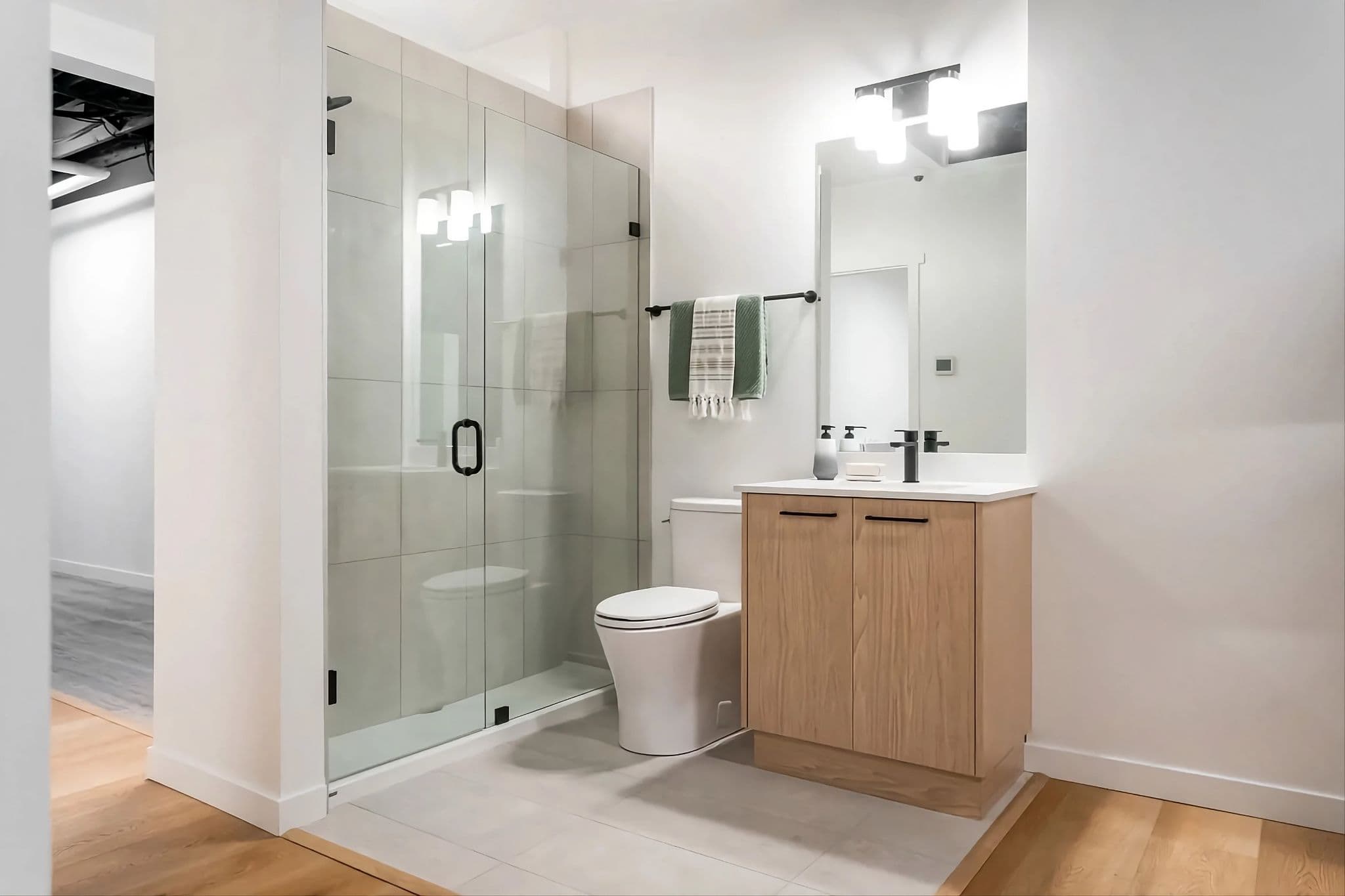 City and Laurel – A bright, modern bathroom featuring a glass-enclosed walk-in shower lined with large light gray tiles and matte black hardware. Beside the shower stands a toilet and a light wood grain vanity with a white countertop, matte black faucet, and a sleek rectangular mirror illuminated by contemporary vanity lighting.