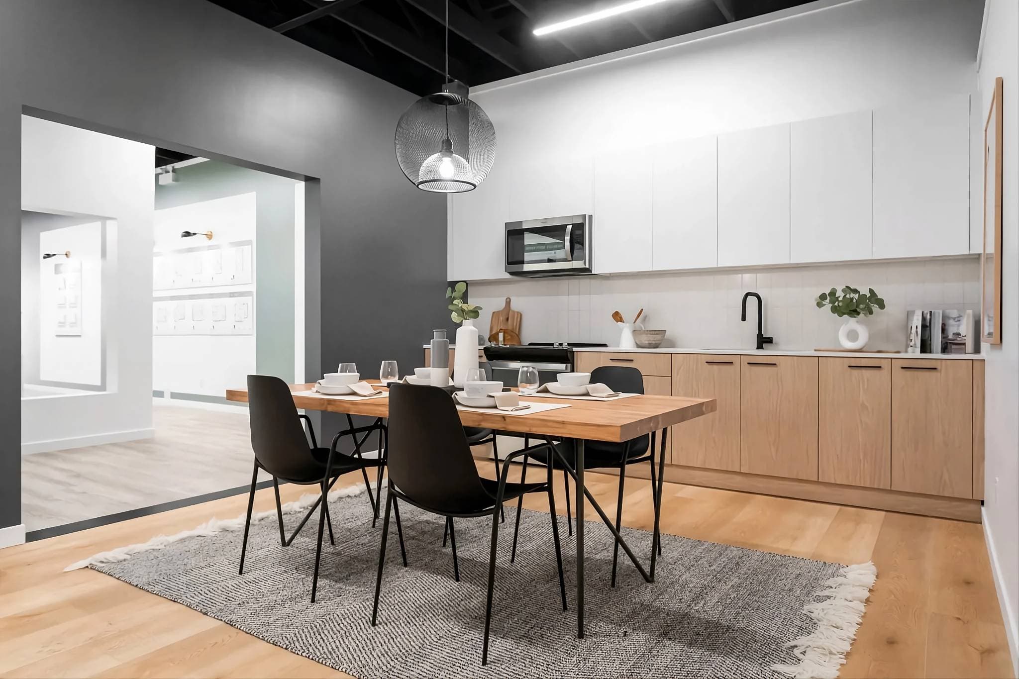 City and Laurel – A modern staged open-concept kitchen and dining area features light wood lower cabinets, flat-panel white upper cabinets, and a set wooden dining table with black minimalist chairs on a textured grey area rug. A distinctive wire mesh pendant light hangs above the table, set against a dark grey accent wall and light hardwood flooring.