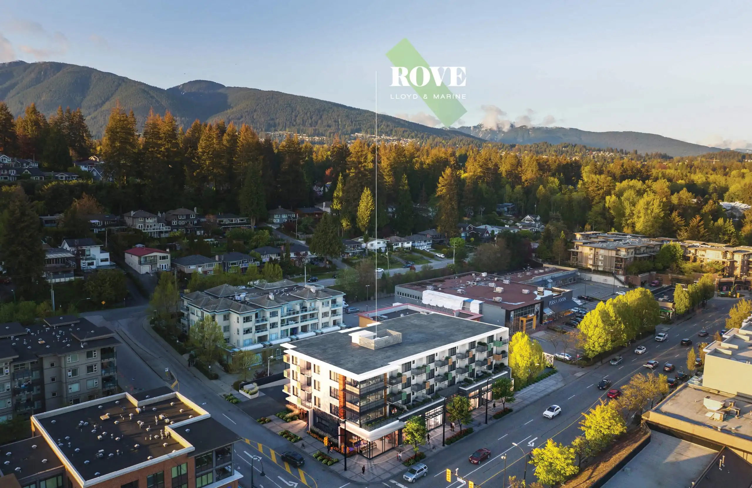 Rove – Aerial drone view of the neighborhood with the building's rooftop marked by a ROVE logo overlay, mountains and forest in the background at sunset.