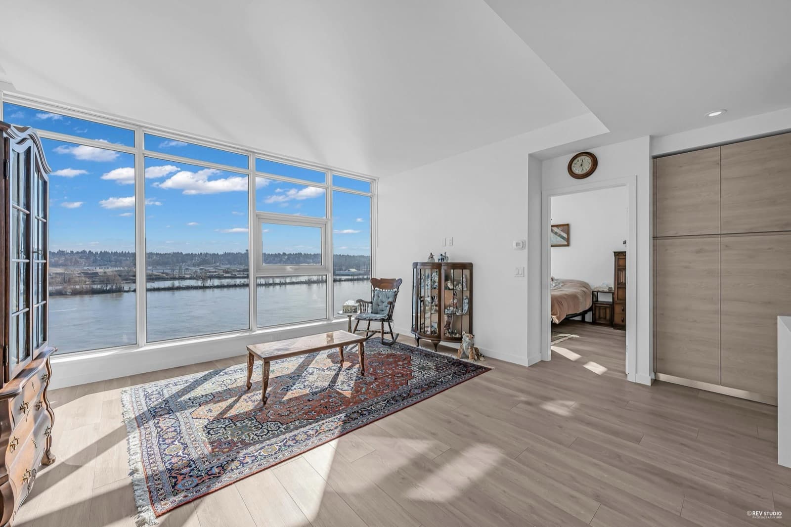 Pier West – Living room with antique furniture, a patterned area rug, and a panoramic window overlooking a river, opening into a bedroom.