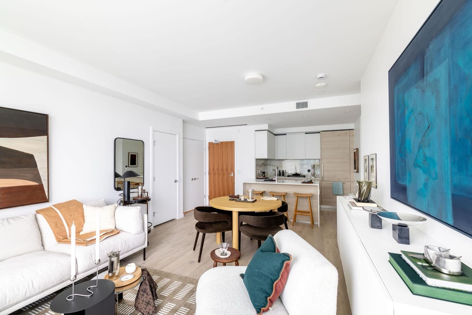 Pier West – Open living, dining, and kitchen area with a blue painting on the wall, a round wood dining table, and dark chairs.