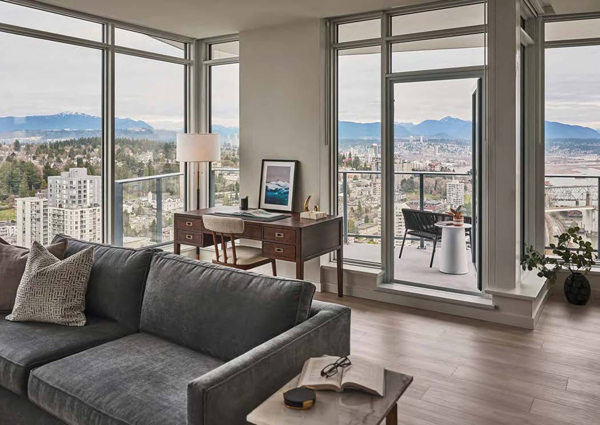 Pier West – Corner living space with a grey sofa, a wood desk and chair, and floor-to-ceiling windows showing mountain and city views plus a balcony.