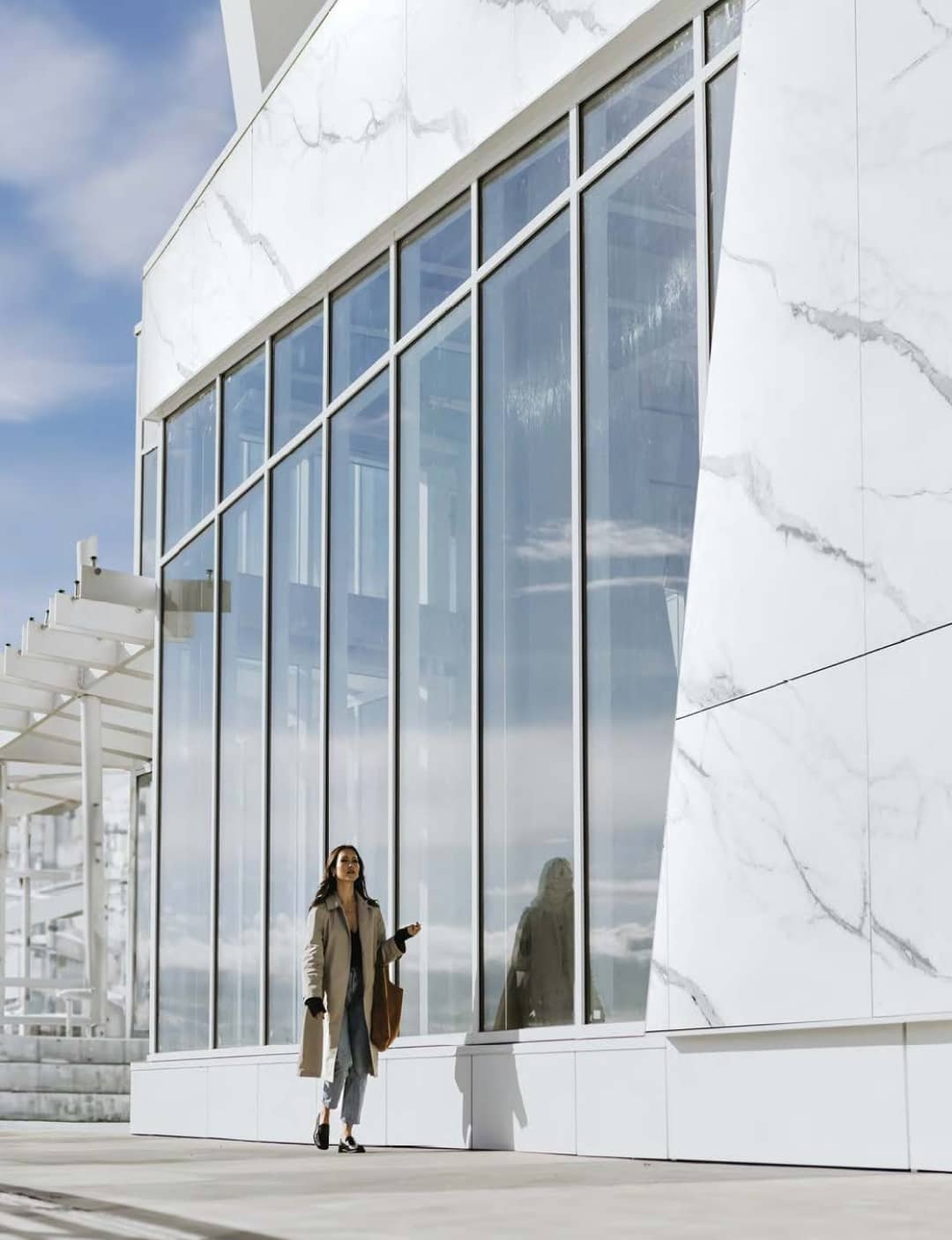 Pier West – Street-level view of the building's white marble-clad facade with tall glass windows, showing a woman walking along the sidewalk.