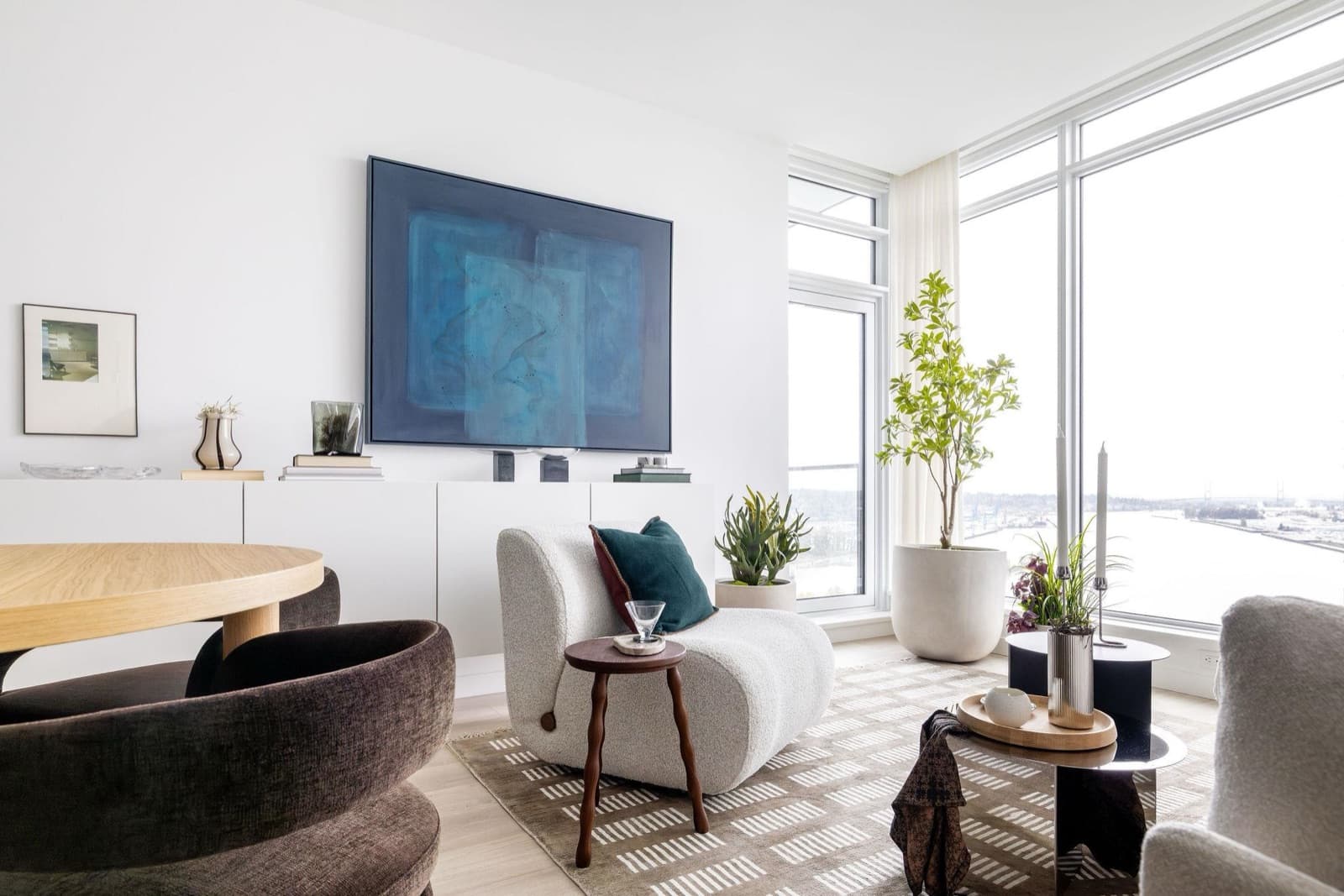 Pier West – Living and dining space with a large blue abstract painting, a cream boucle chaise chair, and floor-to-ceiling windows with a water view.