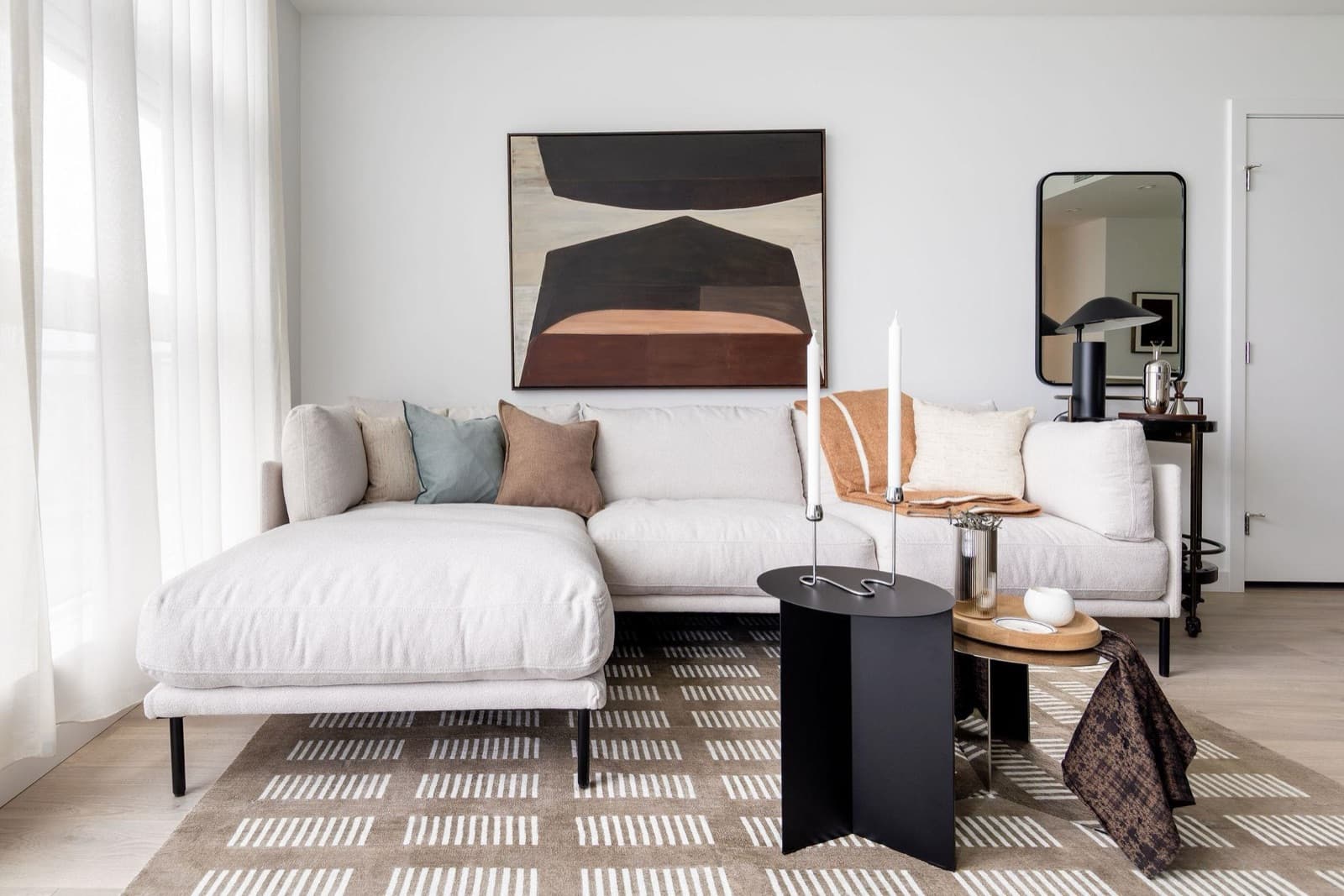 Pier West – Living room with a light grey L-shaped sectional, a dark abstract painting above a credenza, and a patterned area rug.