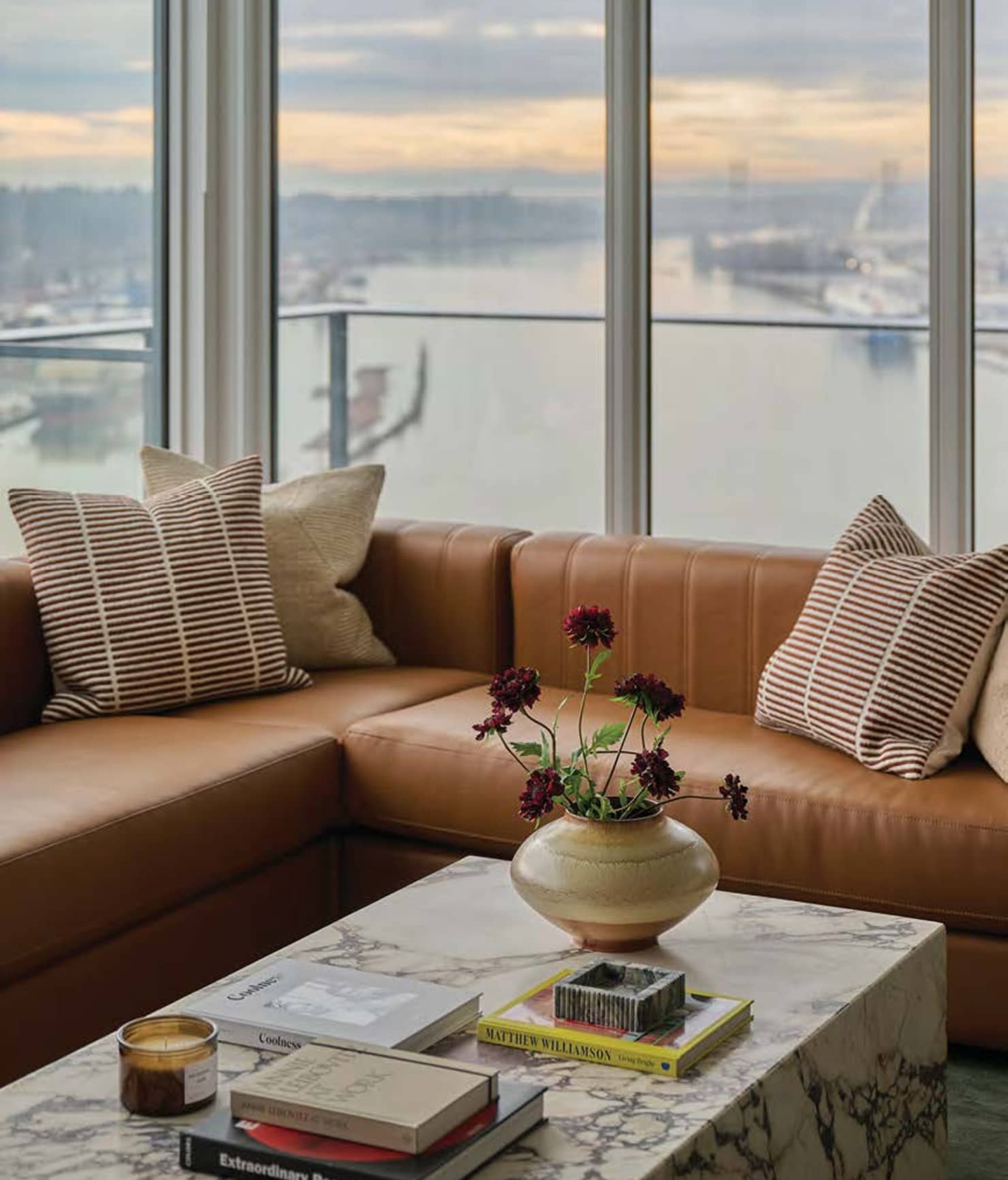 Pier West – Living room with a tan leather sectional sofa, striped throw pillows, and a marble coffee table in front of floor-to-ceiling windows overlooking a river.