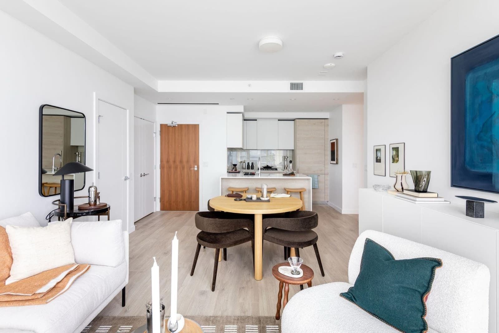 Pier West – Open-concept living and dining area with a round wood table, dark upholstered chairs, and a kitchen visible in the background.