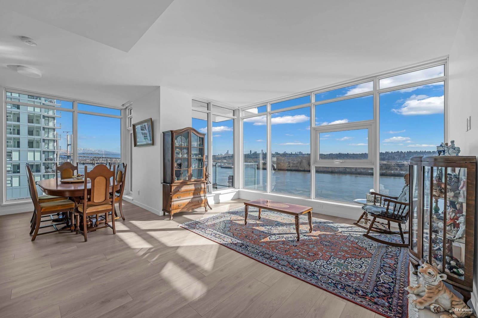 Pier West – Dining and living room furnished with antique wood furniture and an area rug, with floor-to-ceiling windows overlooking a river and a bridge.