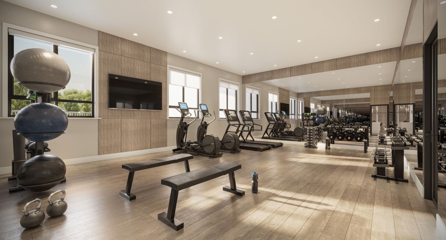 Amson Bloc – A bright, modern residential fitness center equipped with treadmills, stationary bikes, free weights, exercise balls, and workout benches across light wood flooring. Natural daylight streams through large side windows, complementing the warm wood-paneled walls and a full-length mirrored back wall.