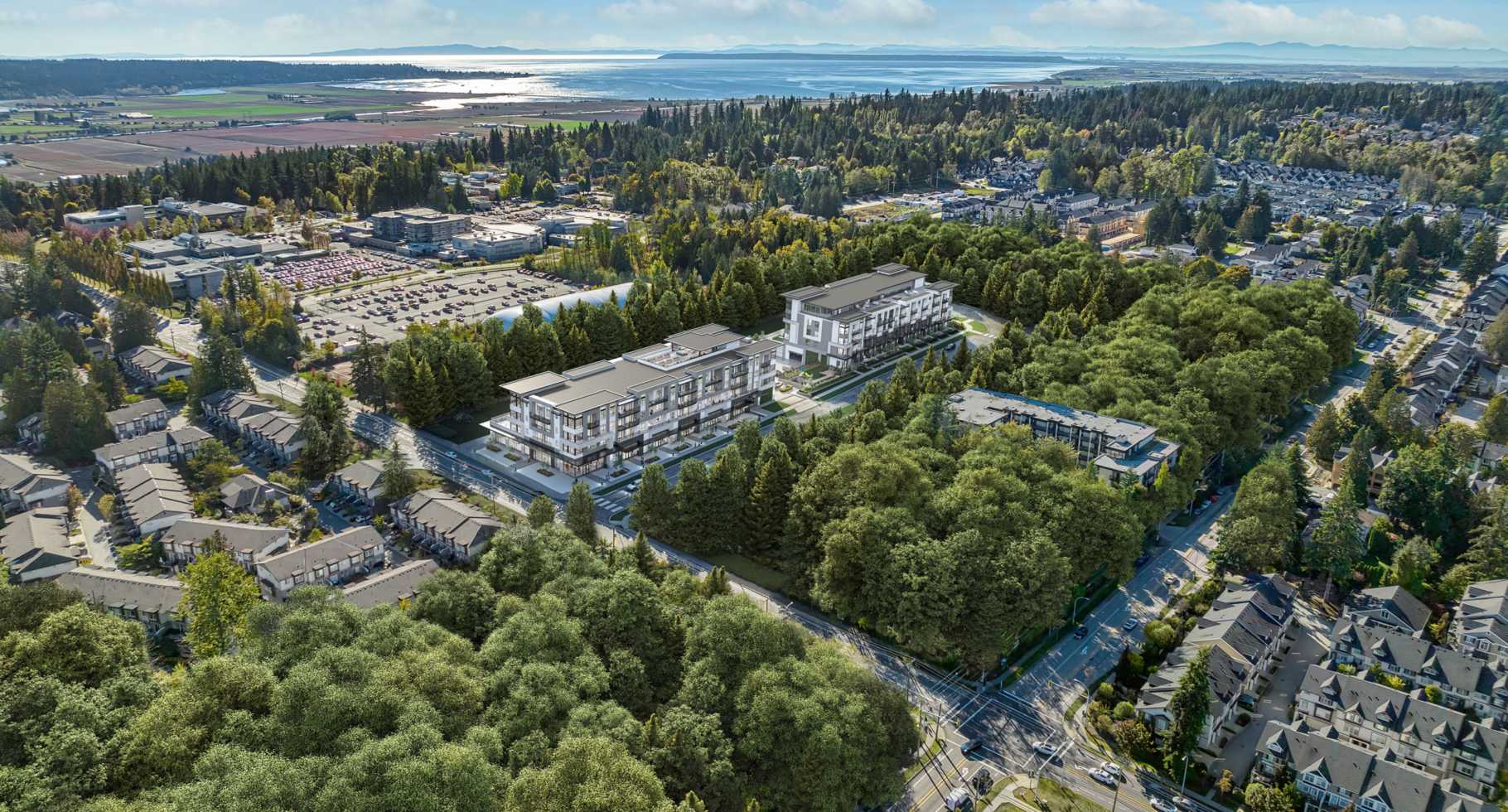 Amson Bloc – A high-angle aerial view depicts contemporary low-rise residential buildings nestled amidst dense green forests and suburban streets under a bright, partly cloudy sky. In the background, the landscape stretches toward open agricultural fields and a wide body of water bordering distant coastal landforms.