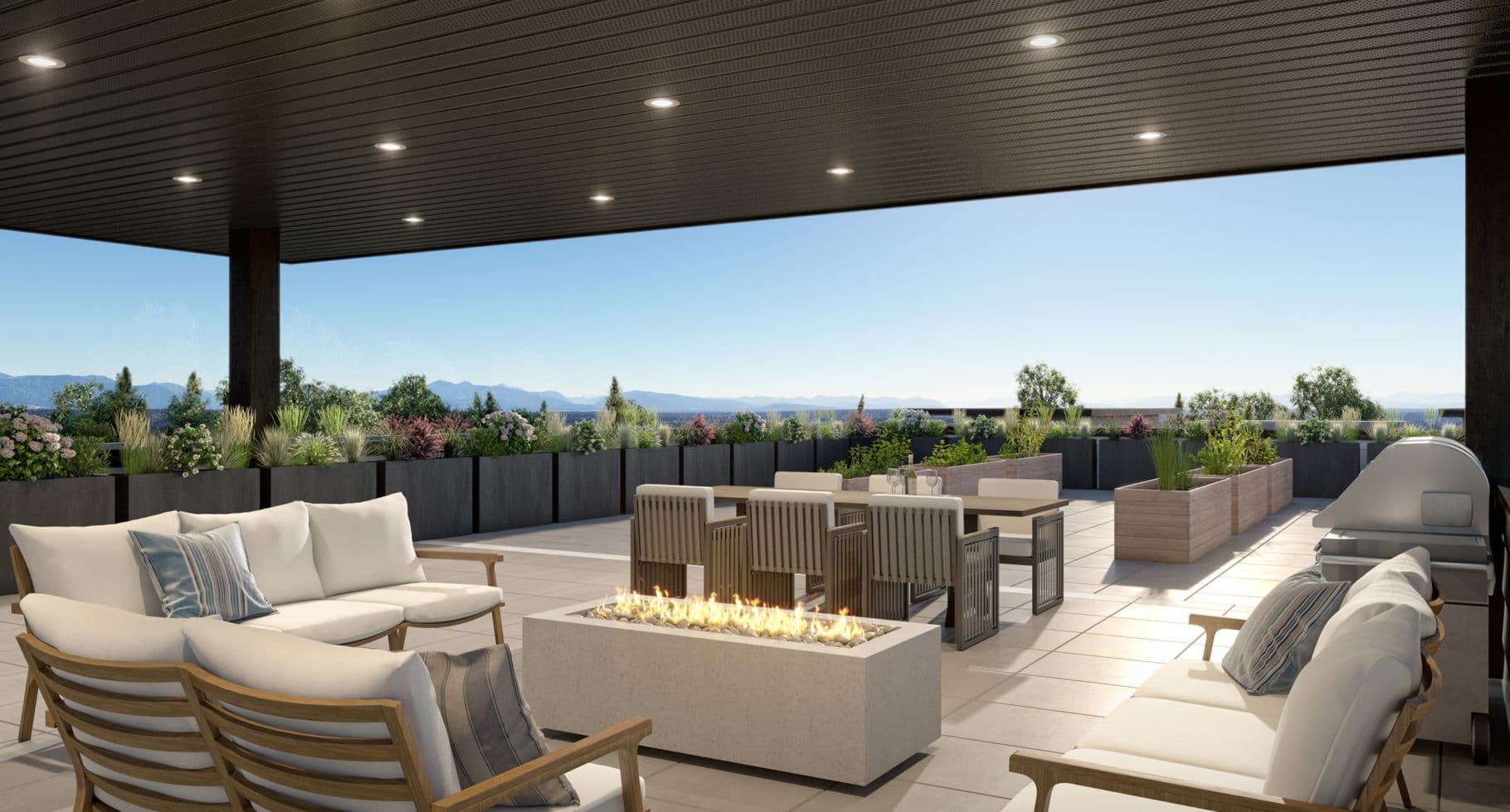 Amson Bloc – A modern outdoor rooftop patio features a rectangular fire pit surrounded by cushioned lounge seating, an al fresco dining table, and a stainless steel BBQ grill. Planter boxes filled with greenery line the edge of the tiled terrace beneath a dark paneled overhang with recessed lighting, looking out over distant mountains on a clear day.
